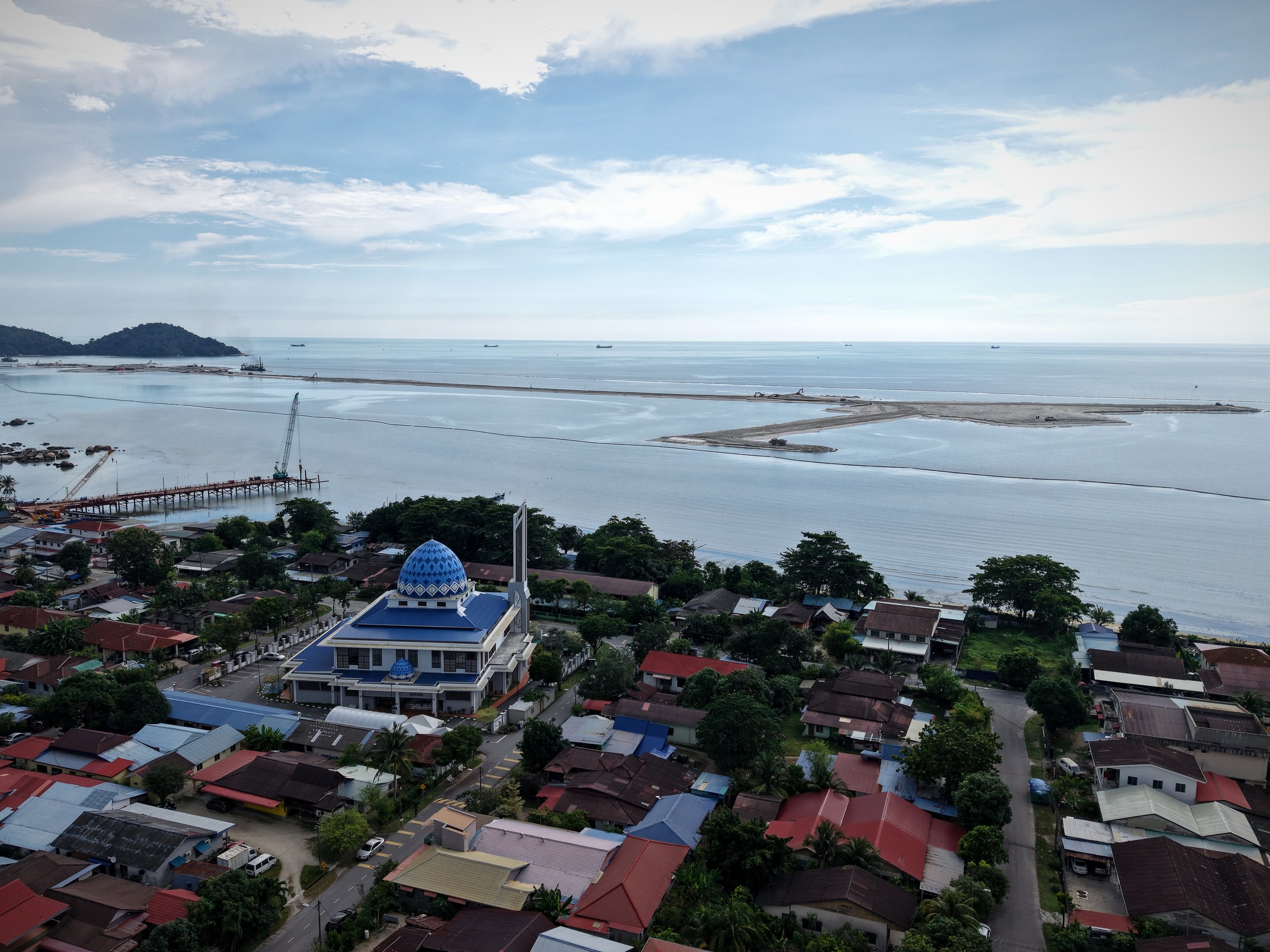 Aerial view over the rooftops of Permatang Damar Laut, a coastal village with a mosque, and the Silicon Island reclamation offshore