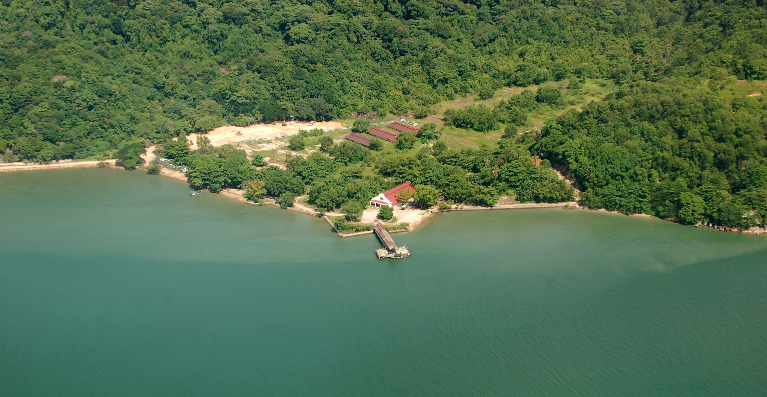 Aerial view of the forested shore of Pulau Jerejak, with a small jetty and a row of red-roofed buildings